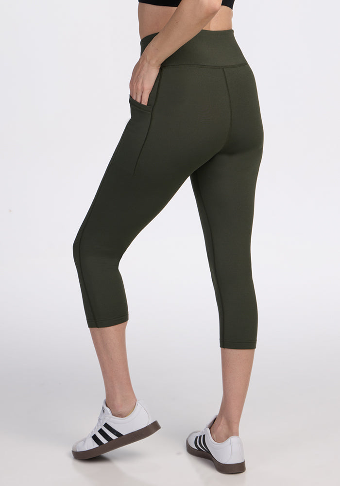 A person wearing Woolx Merino Flex® McKenna Capris in Everleaf and white sneakers with black stripes stands against a plain white background.