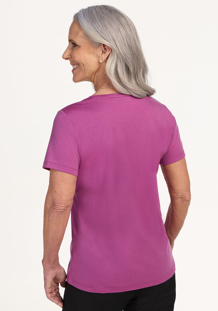 An older woman with gray hair, wearing the Woolx Junie V Neck in Purple Wine and black pants, stands facing away and smiles over her shoulder against a plain light background.