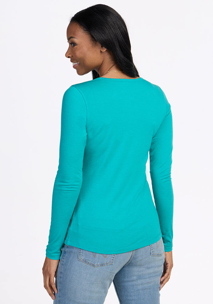 A woman with long dark hair wears the Woolx Remi Long Sleeve T-Shirt in Cool Jade and light blue jeans, standing turned away from the camera and smiling over her shoulder against a plain white background.
