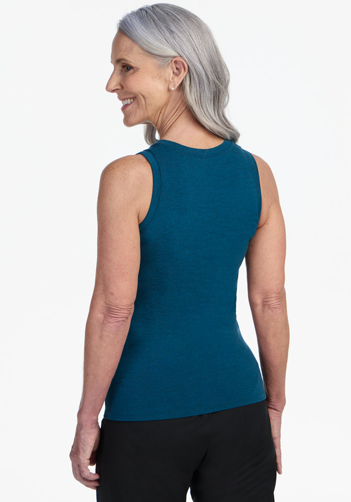 A smiling older woman with gray hair turns slightly to show the back of her Woolx Ava Tank Top in Majolica Blue, paired with black pants, set against a plain white background.