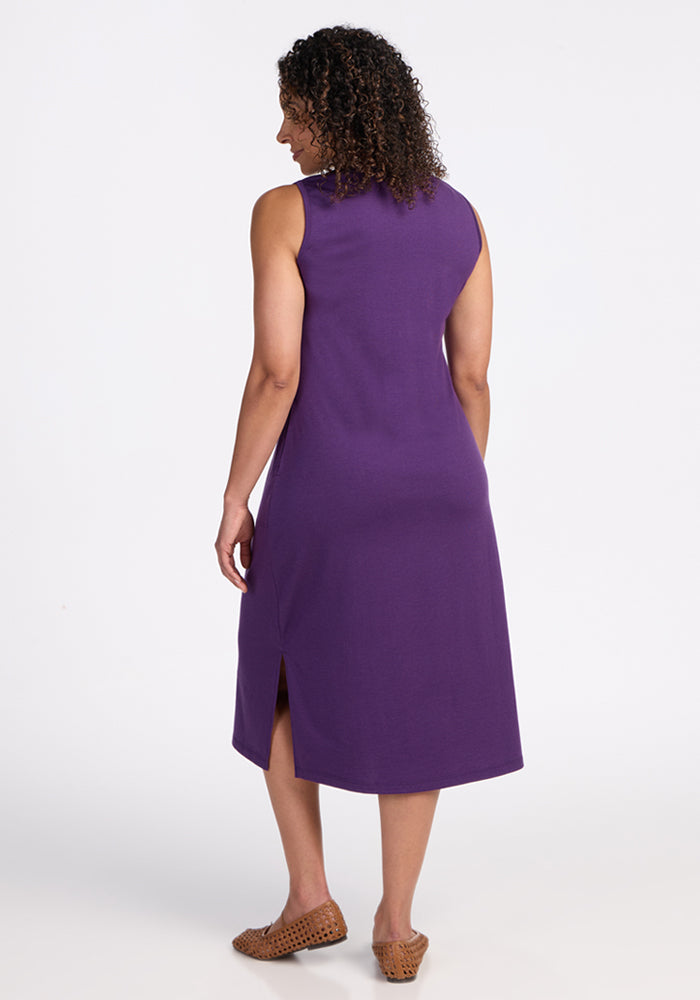 A woman with curly hair faces away, wearing the Woolx Cassie Dress in Plum Crush—a breathable midi dress with a side slit—paired with brown woven flats, against a plain white background.