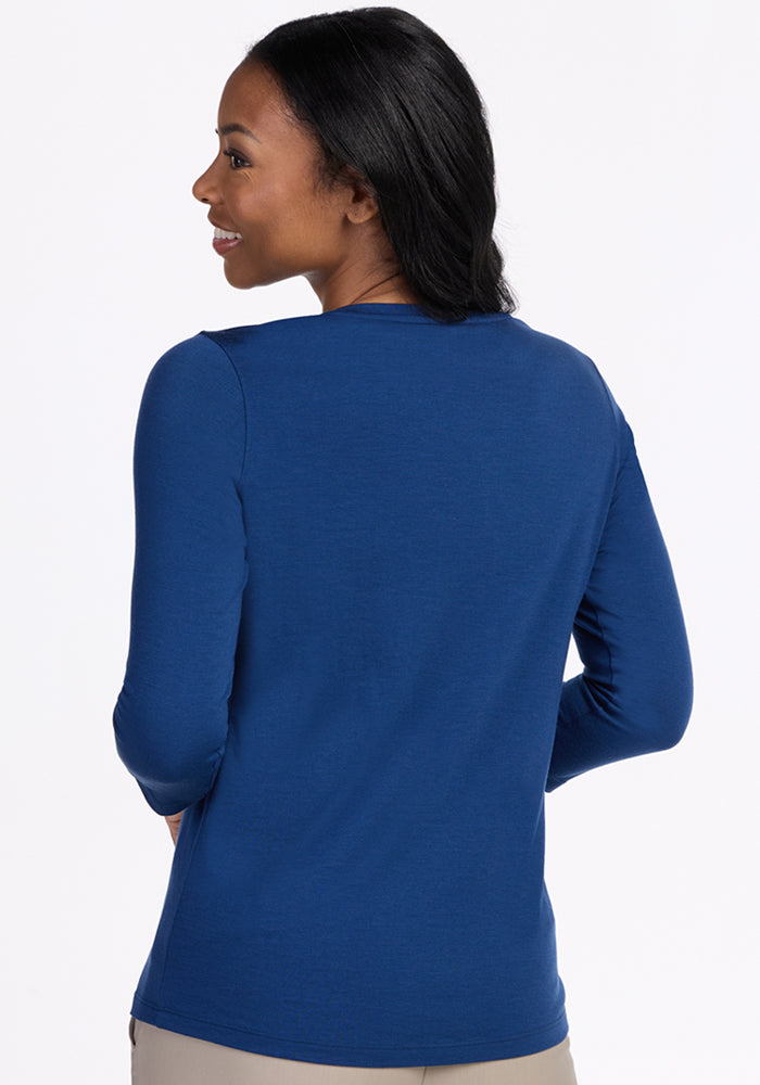 A woman with long dark hair smiles over her shoulder, wearing the Woolx Elena 3/4 Sleeve V Neck in Estate Blue and light-colored pants, set against a plain light background.