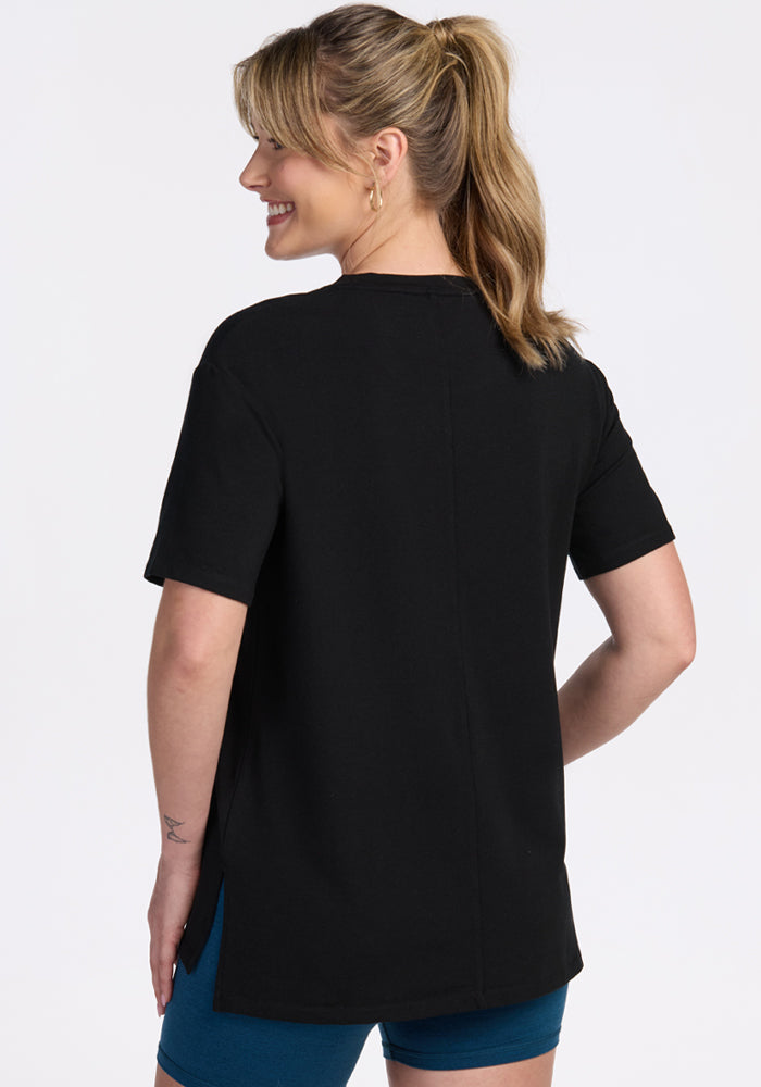 A woman with blonde hair in a ponytail is smiling and facing away from the camera, wearing the Woolx Audrey Tee in black with blue shorts against a plain white background.