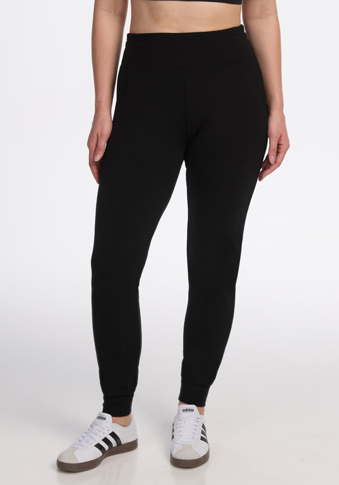 Model wears Woolx Madden Joggers in black, paired with black and white Adidas sneakers and a black crop top, standing against a plain white background. Image shows figure from shoulders down. 