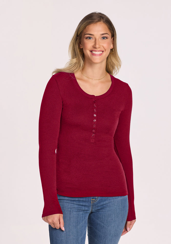 Woman smiles in Woolx Reese Ribbed Henley - Beet Red and blue jeans, standing against a plain light background. 