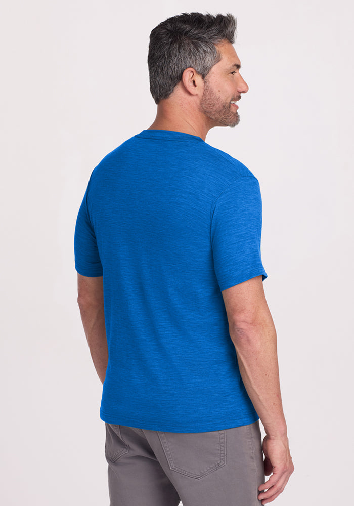 A man with short gray hair and a beard stands facing away, wearing the Woolx Endurance Tee in Glacial Blue and gray pants against a plain light background.