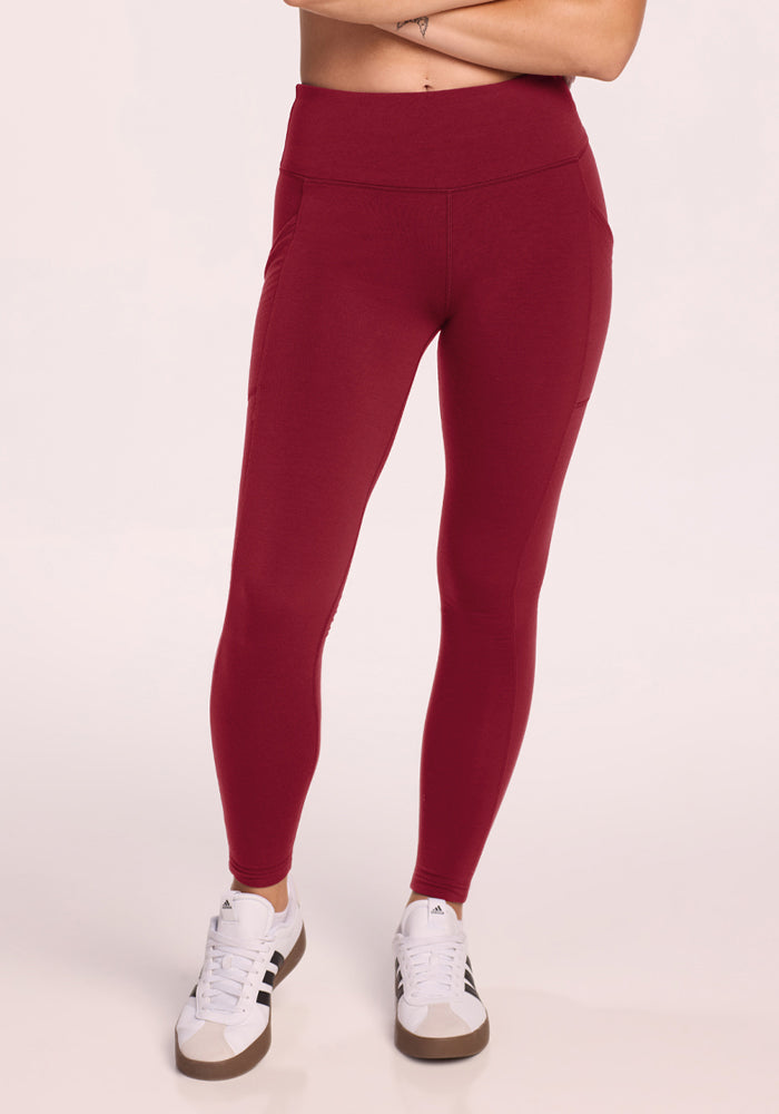 Person wears Woolx Piper Pocket Leggings in Pinot Noir and white sneakers, standing against a light background. 