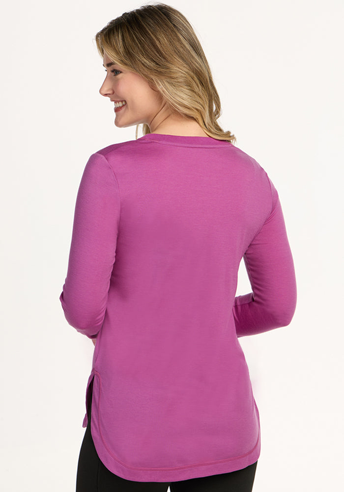 A woman with blonde hair, wearing the Woolx Hazel Tunic in Purple Wine and black pants, stands with her back to the camera and smiles over her shoulder against a plain light background.
