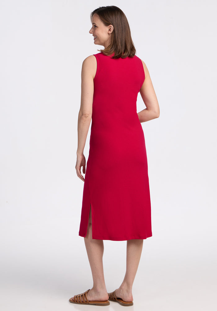 A woman stands smiling, facing away to show the back of the Woolx Iris Ribbed Dress in Poppy Spark. This sleeveless, knee-length merino wool dress with side slits is travel-friendly. She pairs it with flat sandals against a white background.
