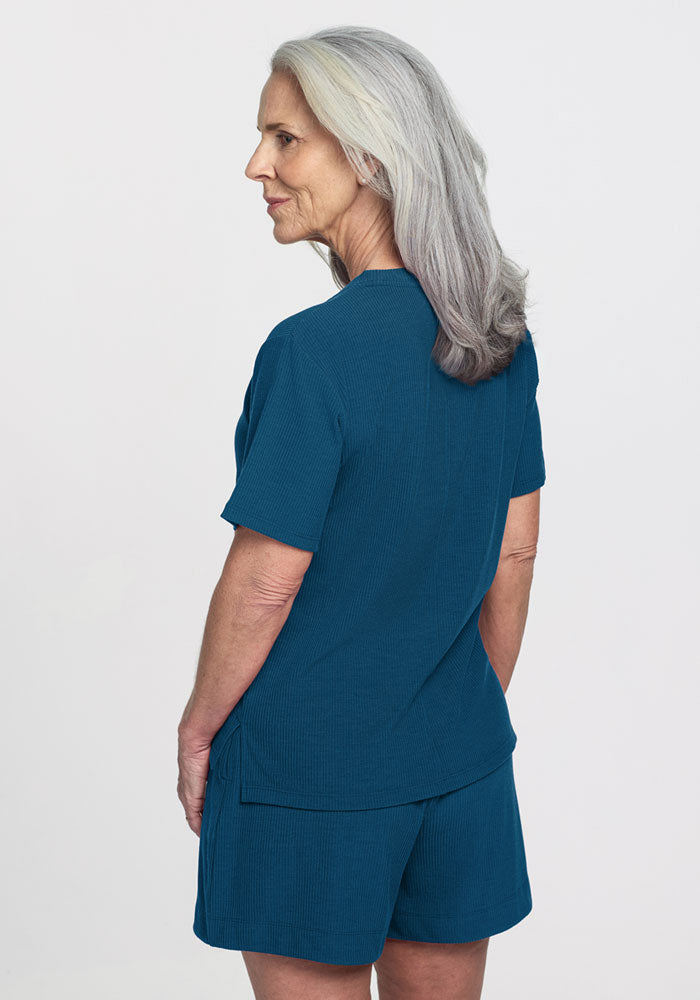 An older woman with long gray hair wears the Woolx Delia Ribbed Short Sleeve top in Moroccan Blue with matching shorts, standing facing away and looking left against a plain white background.