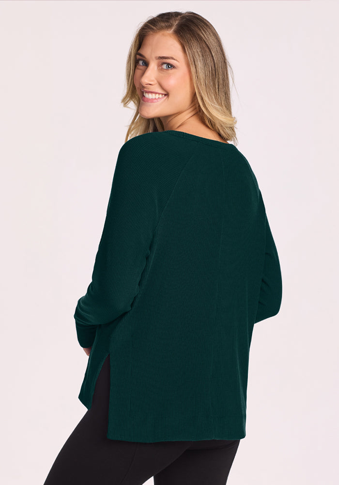 Smiling woman with long hair models the Woolx Ainsley Sweater - Deep Pine, back turned, paired with black pants.
