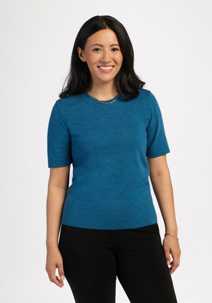 Woman with long dark hair smiles, wearing a Woolx Anna Crew Sweater - Blue Pine and black pants against a white background. 