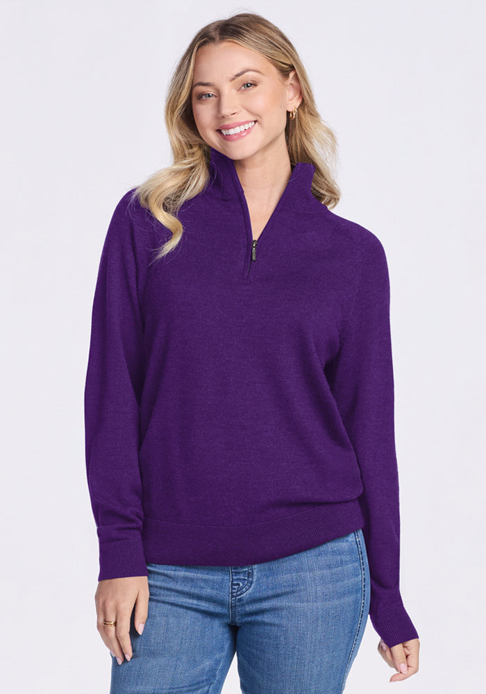 A woman with long, wavy blonde hair smiles while wearing the Woolx Collins Half Zip Sweater in Plum Crush and blue jeans, standing against a plain light background. 