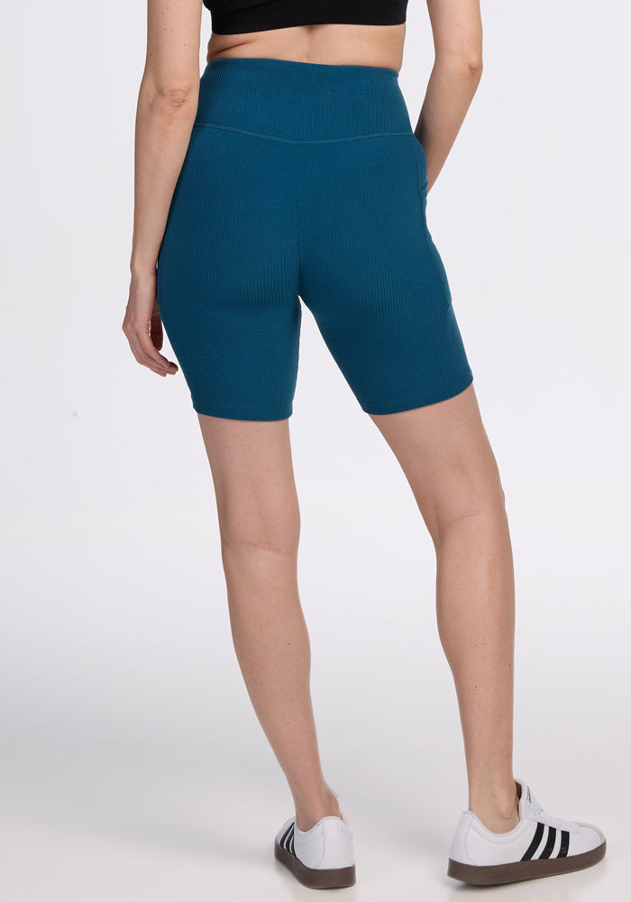 A person stands facing away, wearing a black crop top, Woolx Miley Bike Shorts in Moroccan Blue, and white sneakers with black stripes, against a plain white background.