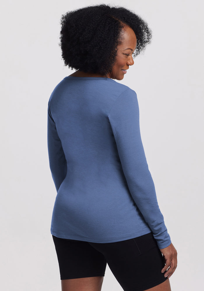 A woman with curly hair wears the Woolx Layla V Neck Top in Skyline and black shorts, standing against a plain light background and smiling over her shoulder.