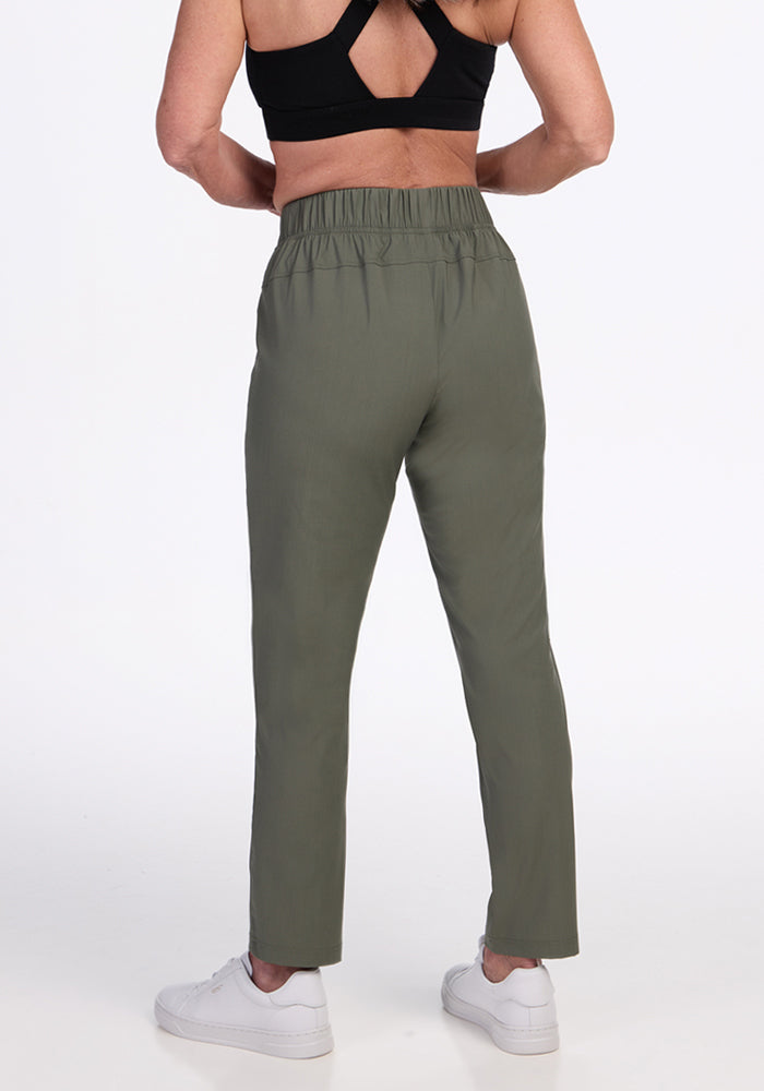 A person stands with their back to the camera, wearing Woolx Nicole Ankle Pants in Sage Olive, a black sports bra, and white sneakers against a plain white background.