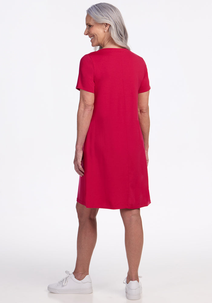 An older woman with long gray hair stands facing away, wearing the Woolx Georgie Dress in Poppy Spark and white sneakers, against a plain white background.