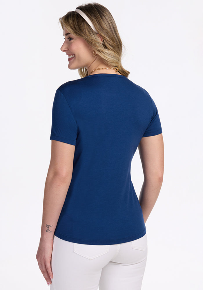 A woman with shoulder-length blonde hair models the Woolx Liza Crew Neck Tee in Estate Blue, paired with white pants. She stands with her back to the camera, smiling over her shoulder against a white background.
