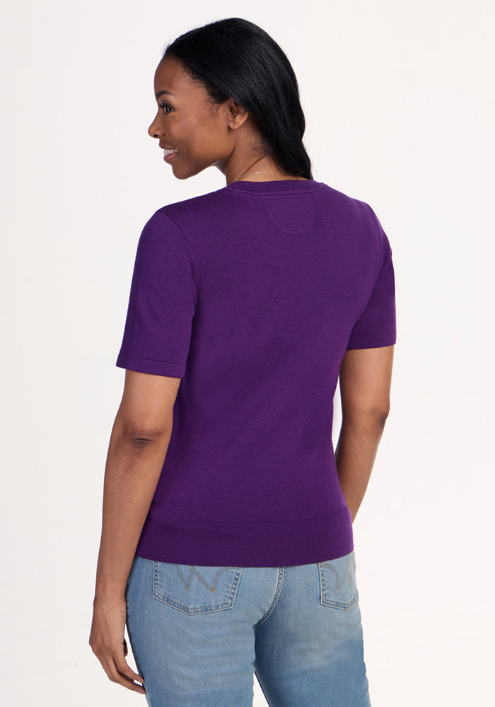 A woman with long dark hair stands facing away and looks right, wearing the Woolx Anna Crew Sweater in Plum Crush and light blue jeans. The background is plain and light-colored.