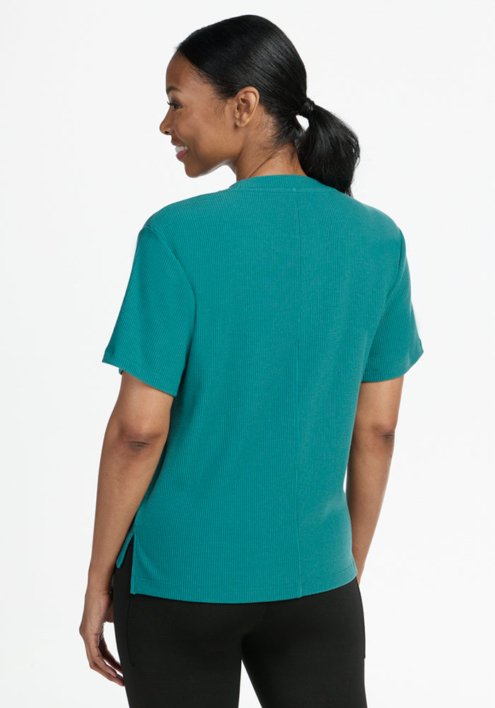A woman with dark hair tied back is shown from behind, modeling the Woolx Delia Ribbed Short Sleeve in Surfside and black pants against a plain white background. She smiles slightly, highlighting the relaxed fit of her outfit.