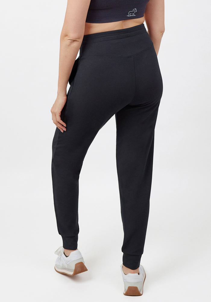 A person wears a black sports bra, Woolx Madden Joggers - Black, and white sneakers while standing with their back to the camera against a plain white background.