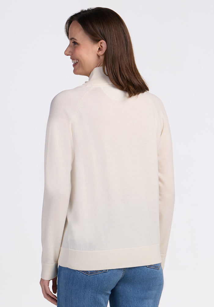 A woman with straight brown hair, wearing the Woolx Collins Half Zip Sweater in Dewdrop Pearl and blue jeans, stands facing away and smiles over her shoulder against a plain white background.