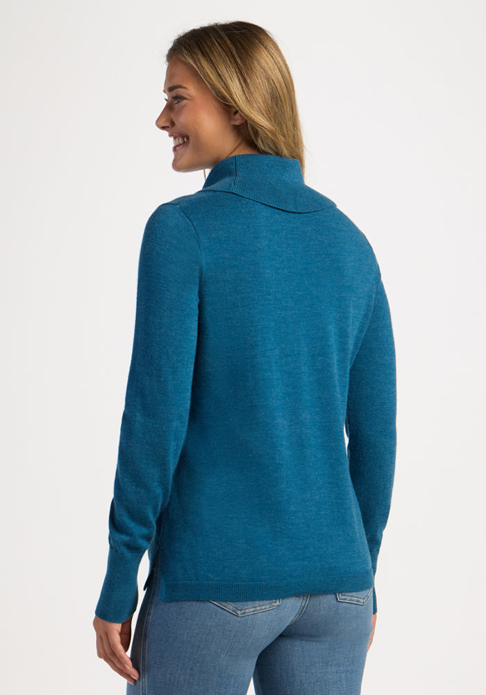 Woman smiles in Woolx Josie Cowl Neck Sweater - Blue Pine, seen from the back, paired with jeans on white background.