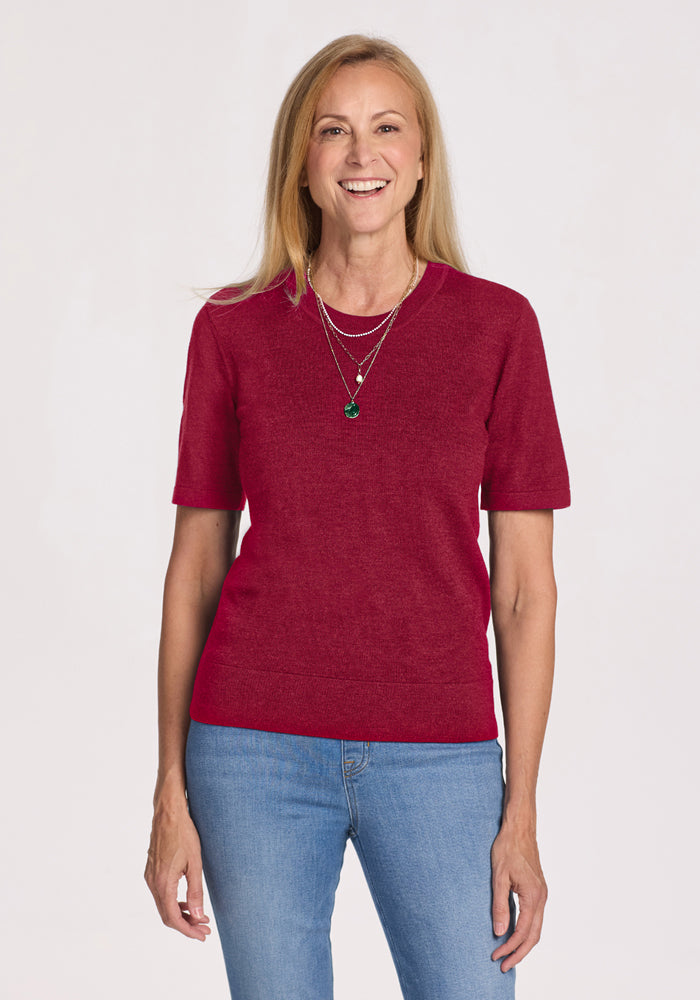Smiling woman with long blonde hair wears a Woolx Anna Crew Sweater in Beet Red, blue jeans, and layered necklaces. 