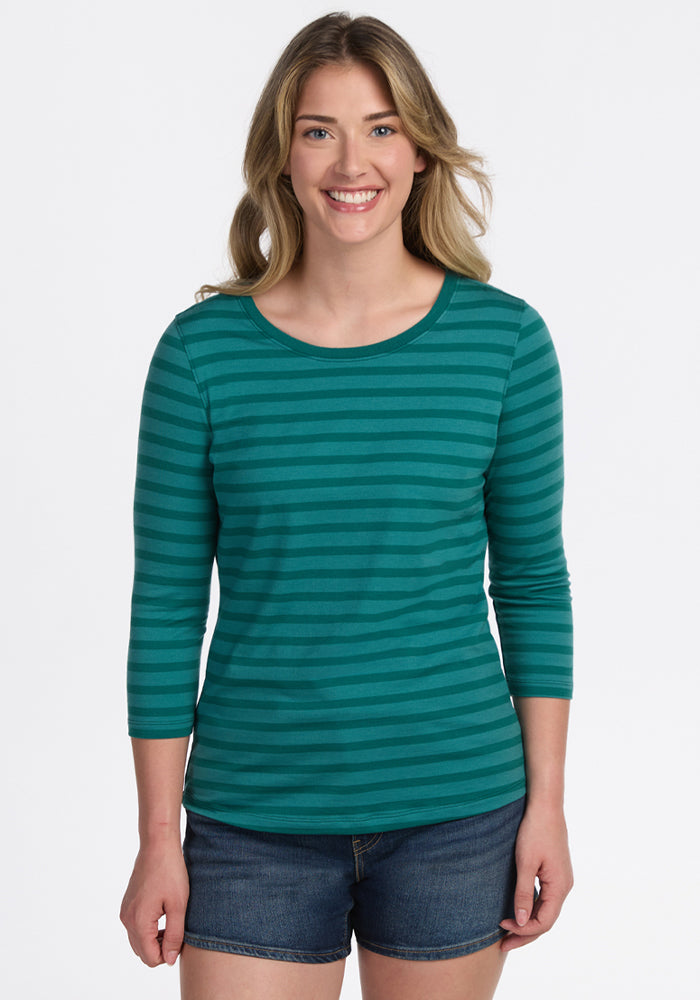 A woman with wavy blonde hair smiles at the camera, wearing the Woolx Jenny 3/4 Sleeve Crew Neck in Woodland Stripe and denim shorts, standing against a plain white background. 