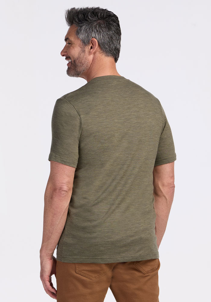 A man with short gray hair and a beard smiles with his back to the camera, wearing the Woolx Endurance Tee in Sage Olive Melange and brown pants as performance gear, set against a plain white background.