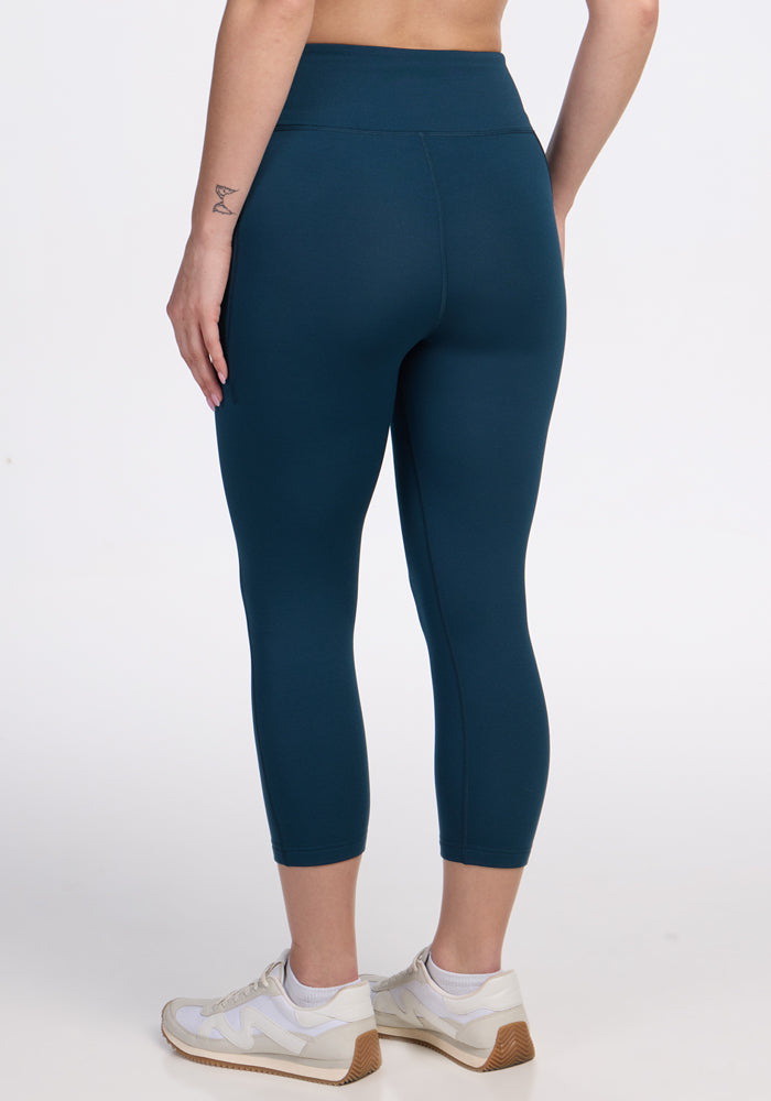 A person wearing Woolx Merino Flex® McKenna Capris in Majolica Blue and white sneakers stands against a plain white background, shown from the back and waist down. A small tattoo is visible on their left forearm.