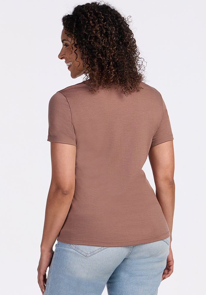 A woman with curly hair stands facing away from the camera, wearing the Woolx Liza Crew Neck Tee in Sienna Kiss and light blue jeans against a plain light background.