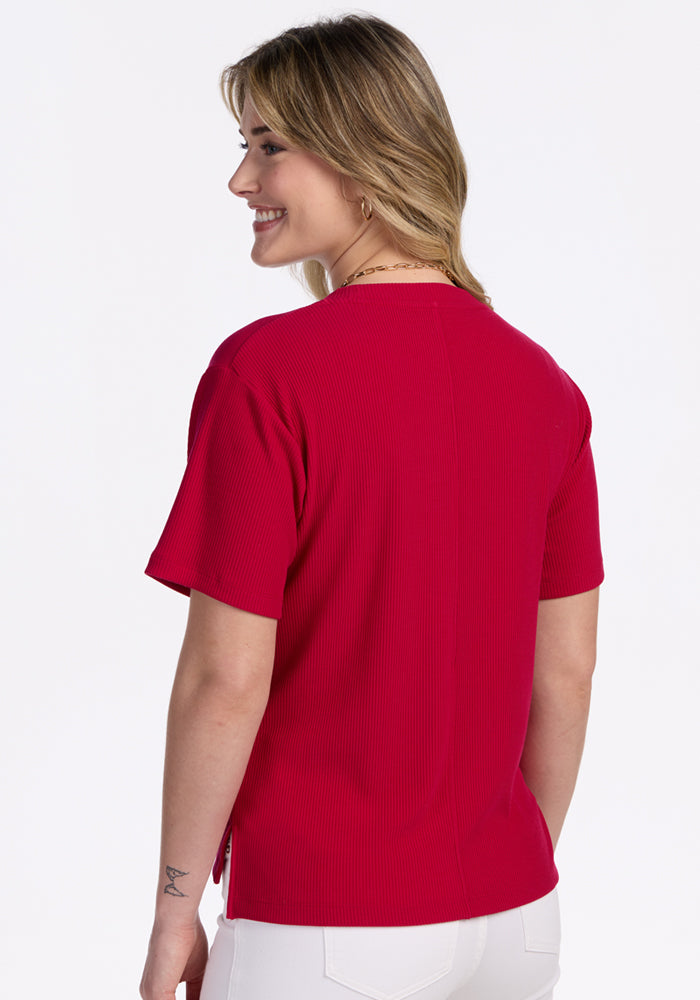 A woman with light brown hair smiles over her shoulder, wearing the Woolx Delia Ribbed Short Sleeve in Poppy Spark with white pants against a plain white background.