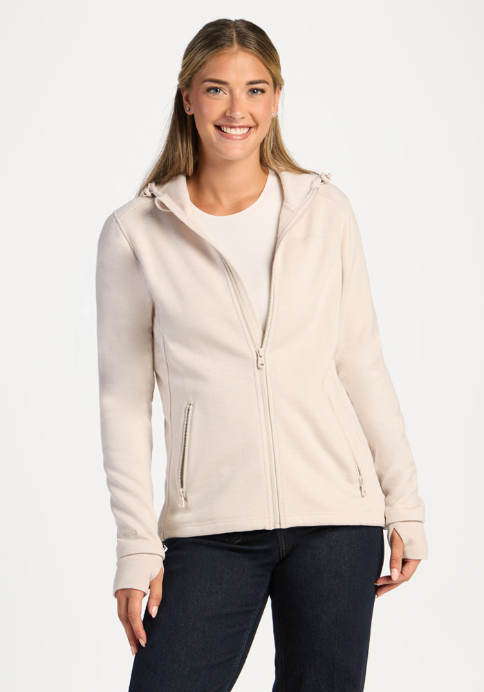 A woman wears an ecru Woolx Cubby Hooded Sweatshirt, a warm fleece zip-up with thumbhole cuffs and zippered pockets. 