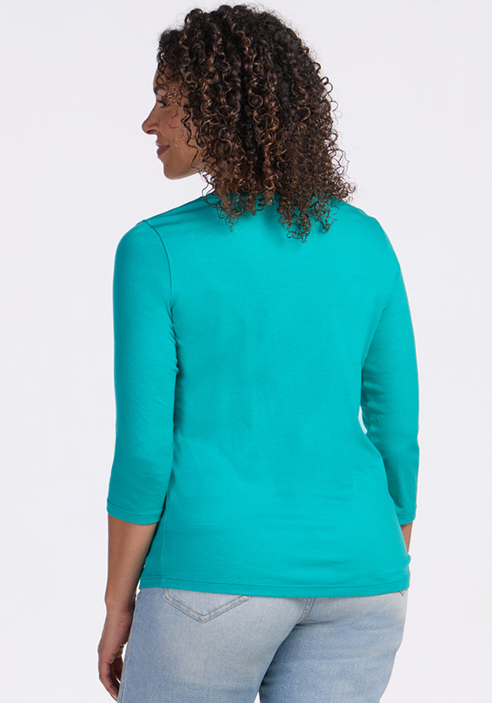 A woman with curly hair, wearing the Woolx Elena 3/4 Sleeve V Neck in Cool Jade and light blue jeans, stands with her back to the camera against a plain white background.