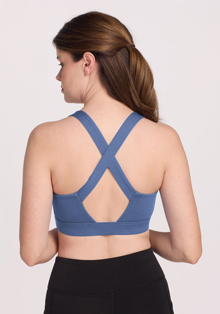 A woman with long brown hair wears the Woolx Livi Bra - Skyline, a blue moisture-wicking sports bra featuring crisscross back straps, paired with black leggings and standing against a plain light background.