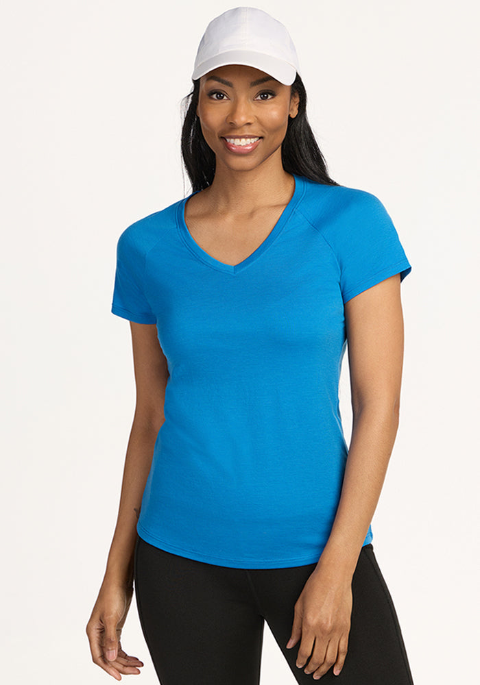 A woman, smiling at the camera against a plain light background, wears the Woolx Mia V Neck - Wave Rider t-shirt, black pants, and a white baseball cap. 