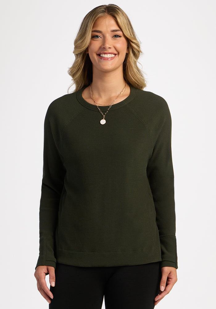 Woman smiles in Woolx Ainsley Petite Sweater—Duffel Bag, dark green, long sleeves, black pants, white background.