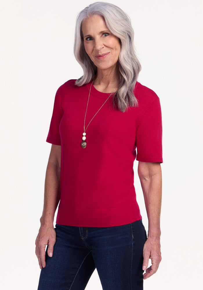 A smiling older woman with long gray hair wears the Woolx Anna Crew Sweater in Poppy Spark, dark jeans, and a long necklace, standing against a plain light background. 