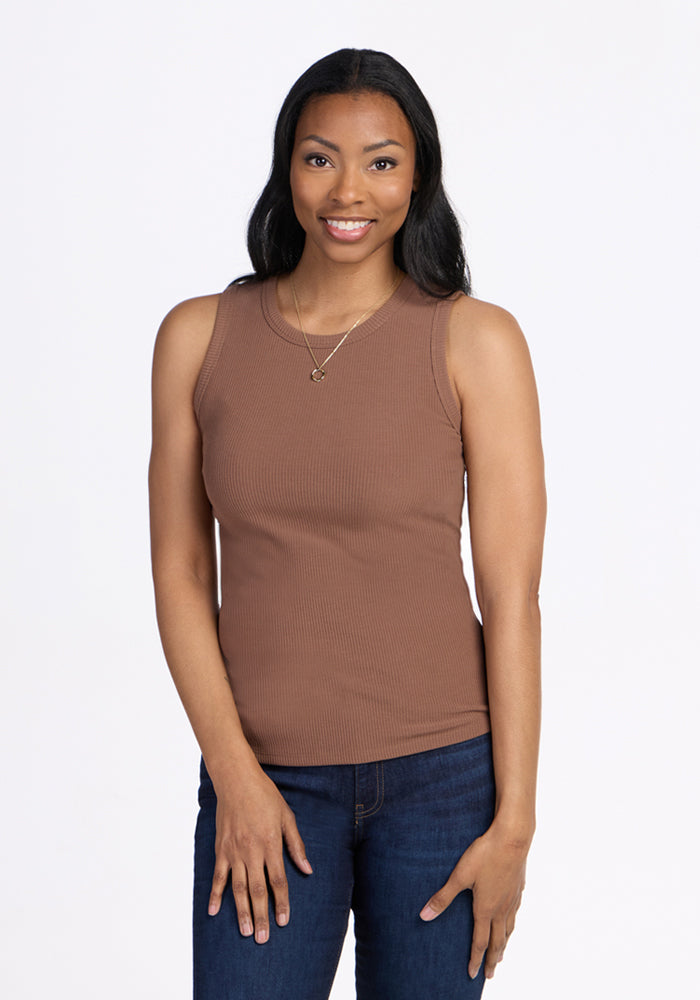 A woman with long dark hair smiles against a white background, wearing the Woolx Raegan Ribbed Tank Top in Sienna Kiss and blue jeans. She rests her left hand on her thigh and accessorizes with a necklace. 