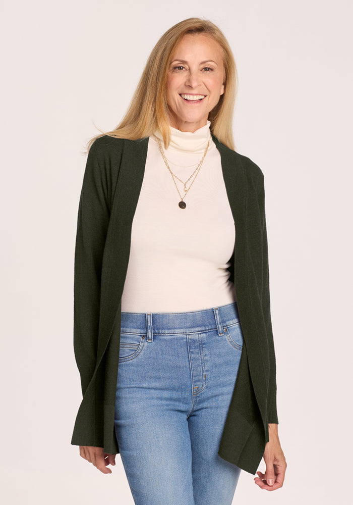 Smiling woman in the Woolx Macie Sweater - Duffel Bag, white turtleneck, and jeans stands against a plain background. 