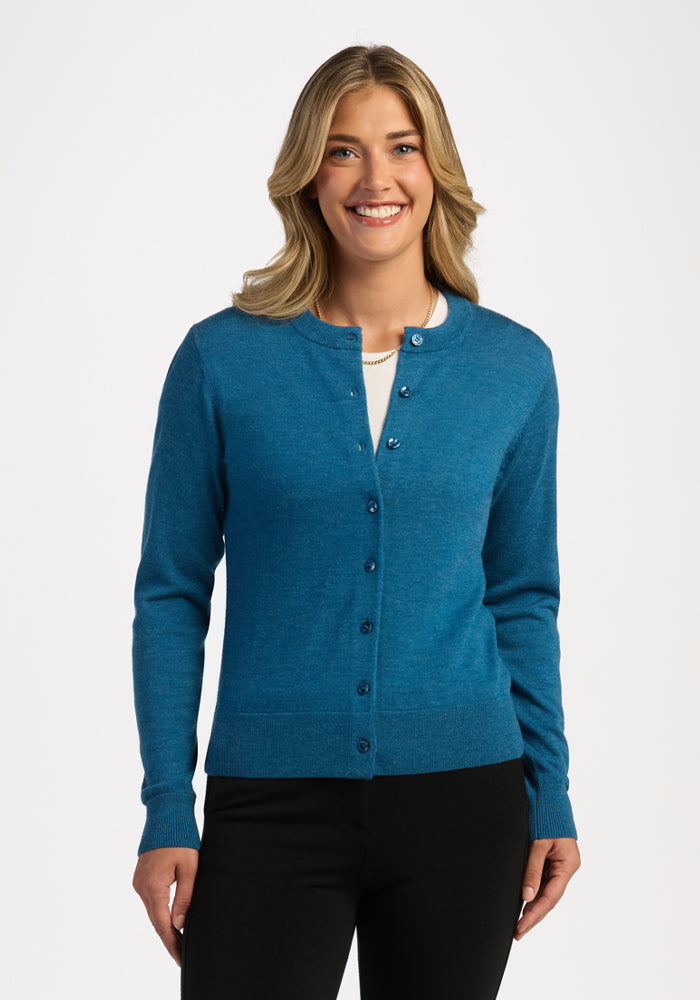 Woman smiling in a Woolx Caitlin Sweater in Blue Pine over a white shirt and black pants, against a white background. 