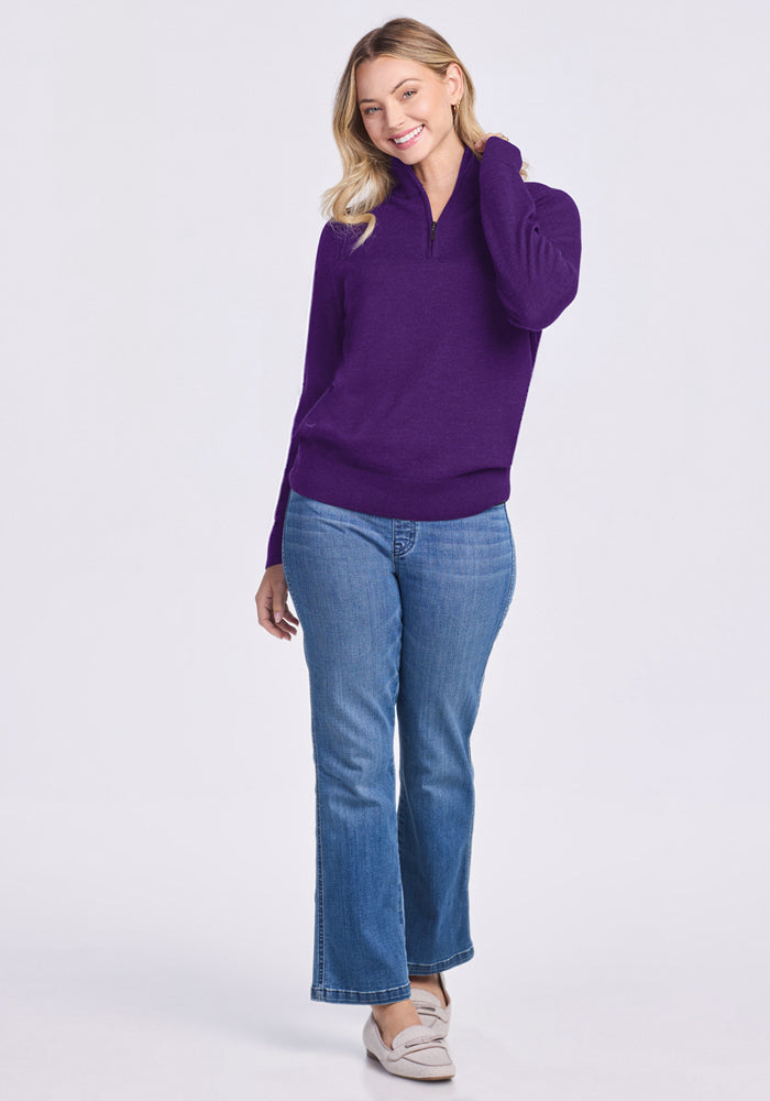 A blonde woman smiles while standing against a plain light background, wearing the Woolx Collins Half Zip Sweater in Plum Crush with blue jeans and beige shoes.