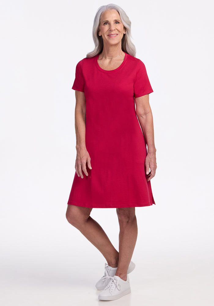 A smiling older woman with long gray hair wears the Woolx Georgie Dress in Poppy Spark and white sneakers, standing on a white background.