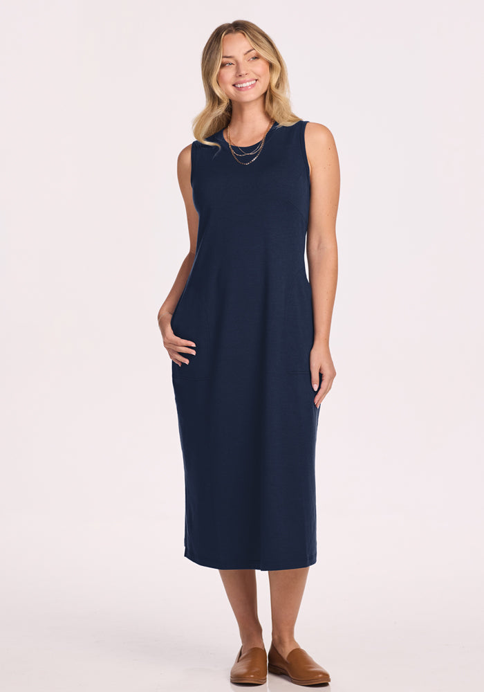 A woman with blonde hair wears the Woolx Cassie Dress in Deep Navy, a sleeveless, wrinkle-resistant travel dress, along with brown flats. She smiles with one hand in her pocket against a plain light background. | Saige is 5’7”, wearing a size XS
