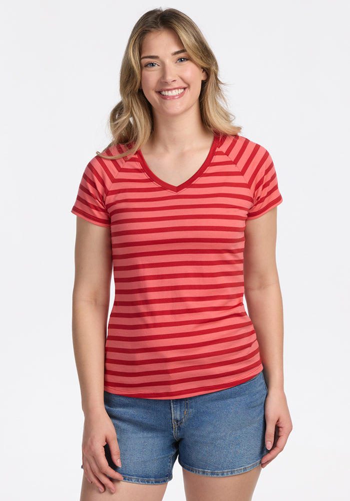 A smiling woman with long, light brown hair wears the Woolx Mia V Neck in Wildberry Stripe, paired with blue denim shorts, standing against a plain light background. 
