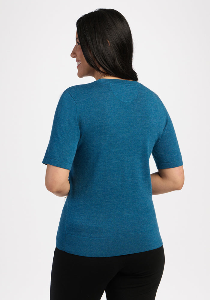 Woman in a blue pine Anna Crew Sweater by Woolx and black pants, standing with back to camera on white background.