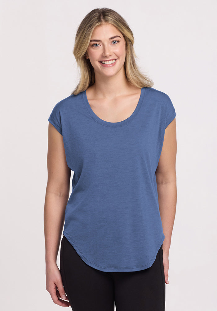 A smiling person with long blonde hair is wearing the Woolx Gabi Tunic Top in Skyline and black pants, standing against a plain light background. 