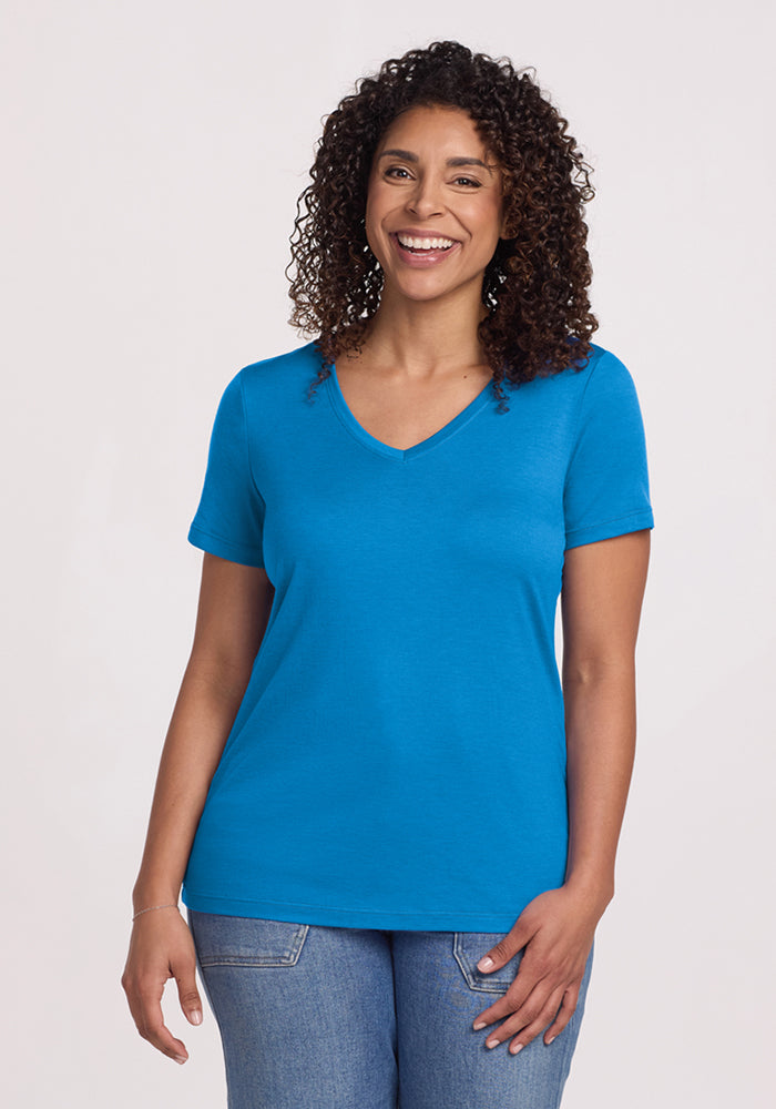 A woman with curly hair smiles against a light background, wearing the Woolx Junie V Neck - Wave Rider and light blue jeans. 