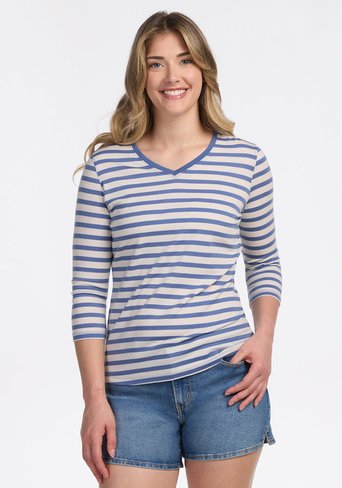 A woman with long, wavy hair smiles at the camera, wearing the Woolx Elena 3/4 Sleeve V Neck in Blue Sand Stripe, paired with denim shorts against a plain white background. 